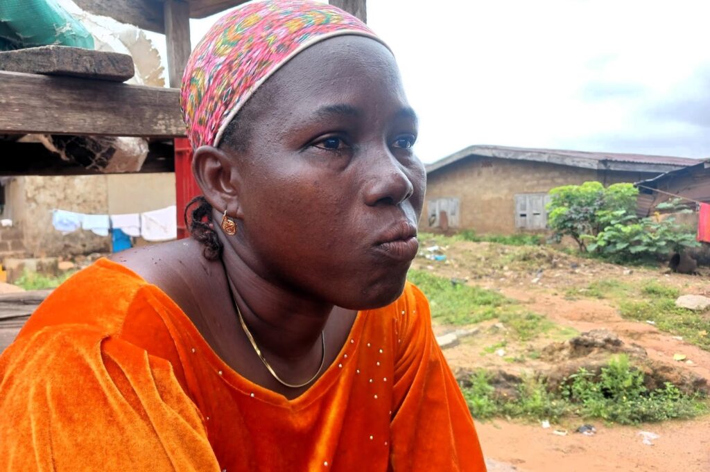 Woman in an orange dress with a headscarf looking pensively to the side, outdoors near a house.