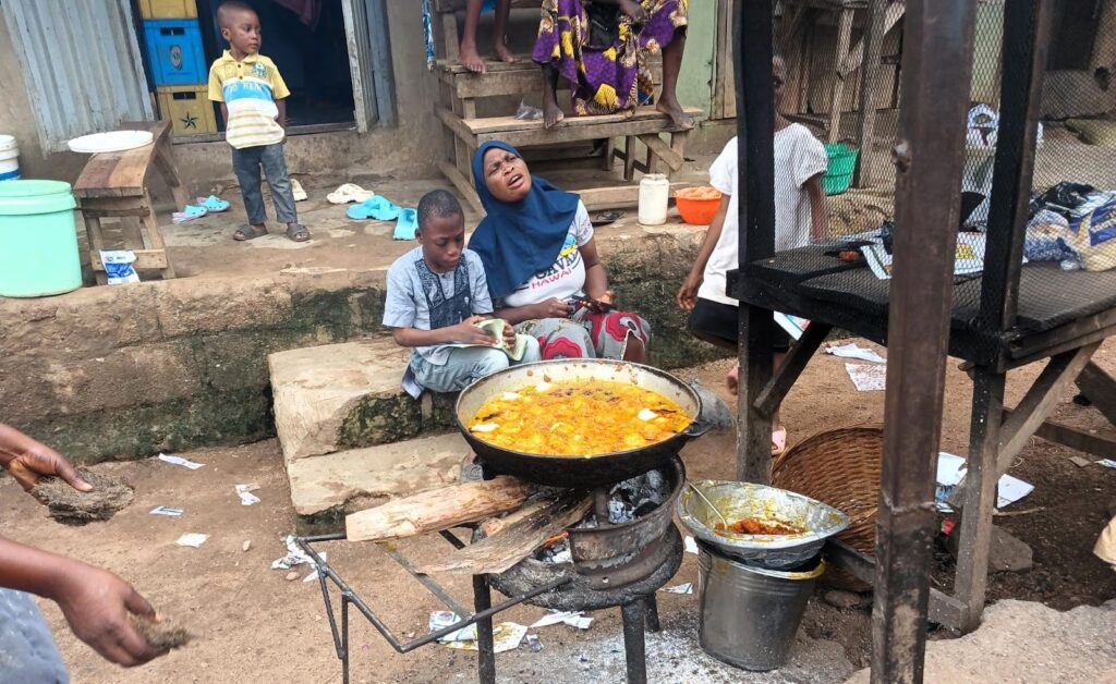 Woman and child near a large frying pan with food at an outdoor street kitchen, with other people nearby.