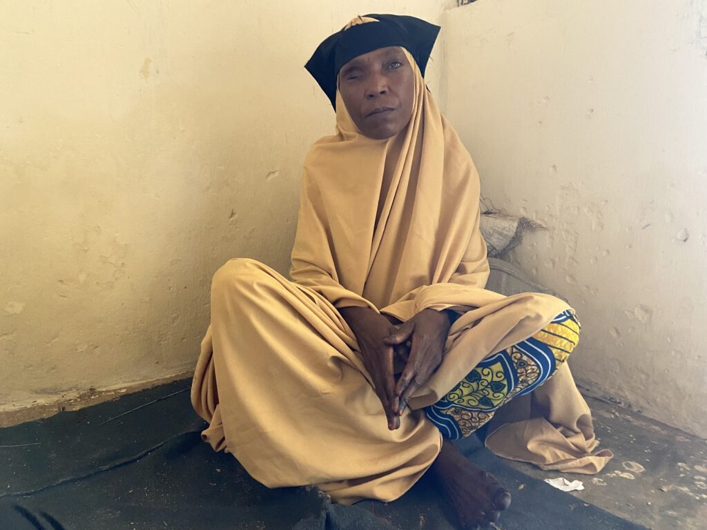 Woman in beige headscarf and robe sitting against a wall.