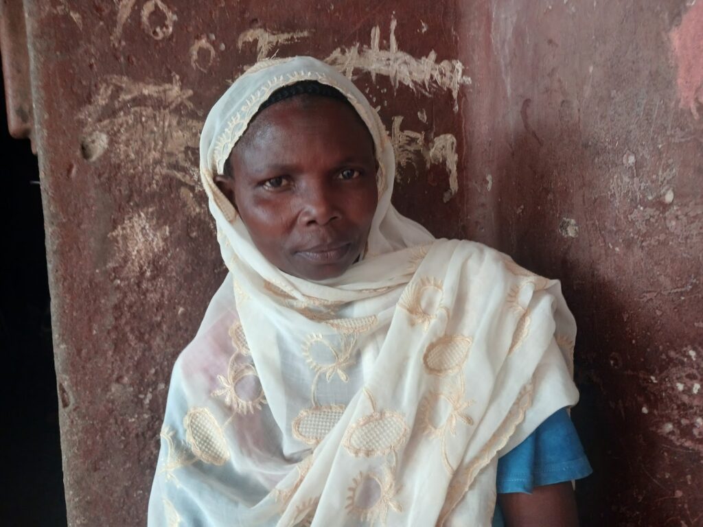 A woman wearing a headscarf standing against a weathered wall.