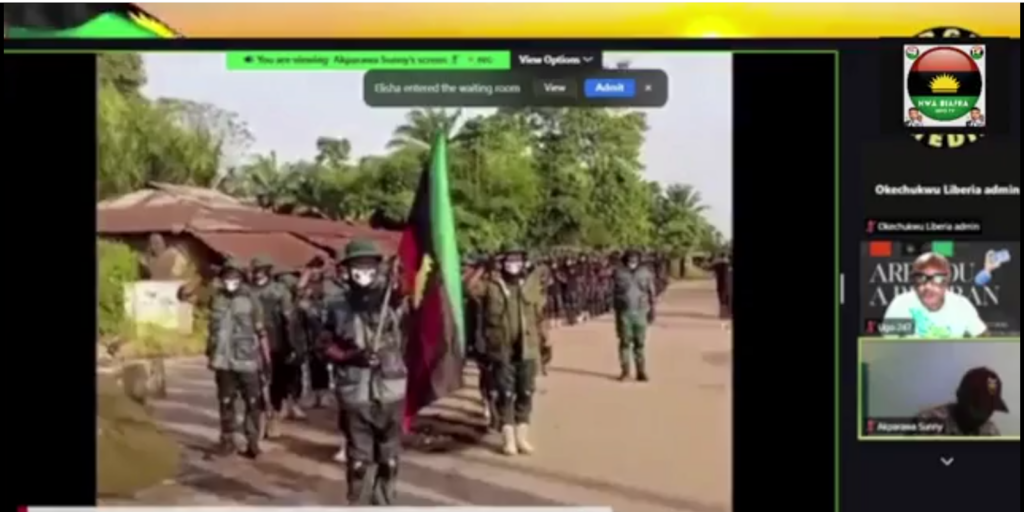 A group of individuals in military attire marching on a road with a flag.