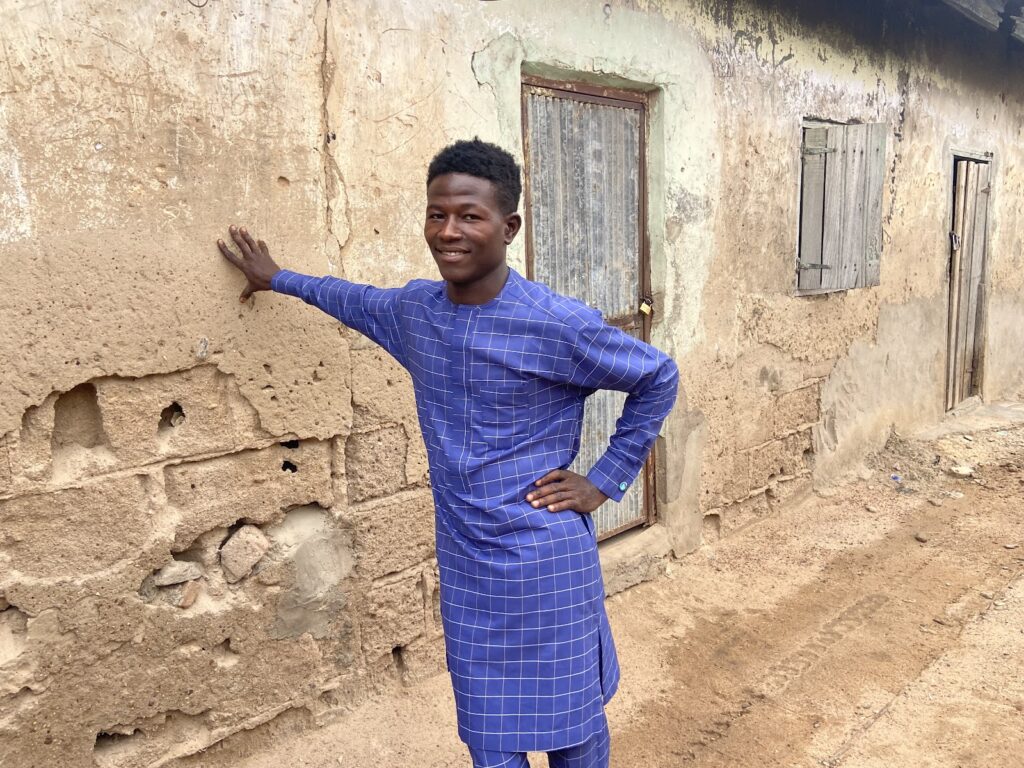 Smiling man in blue attire leaning against a weathered wall with wooden windows.