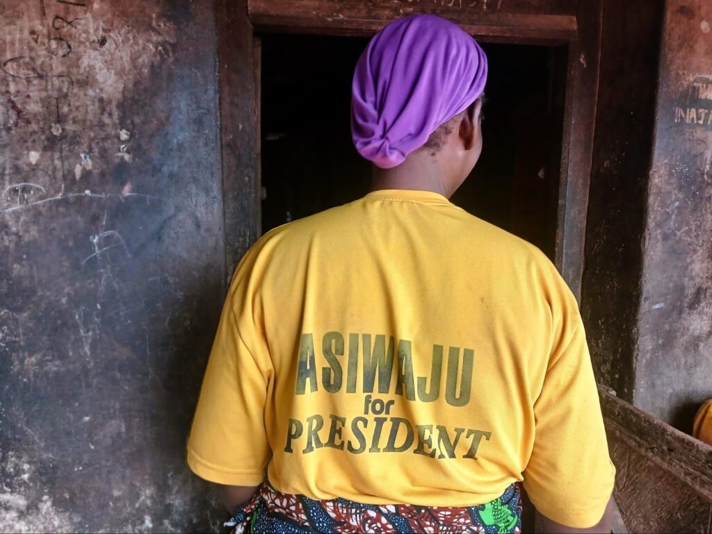 Person in a yellow shirt with "ASIWAJU for PRESIDENT" on the back, standing in a doorway.