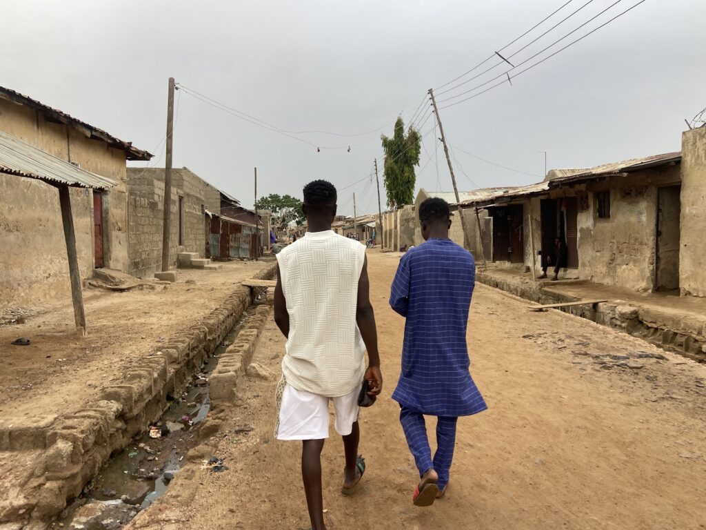 Two men walking down a dirt road in a rural village with houses and a power line overhead.