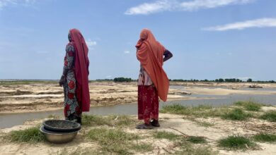 Two women in traditional attire stand by a riverbank looking out over sandy terrain and water.