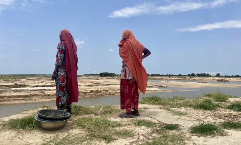 Two women in traditional attire stand by a riverbank looking out over sandy terrain and water.