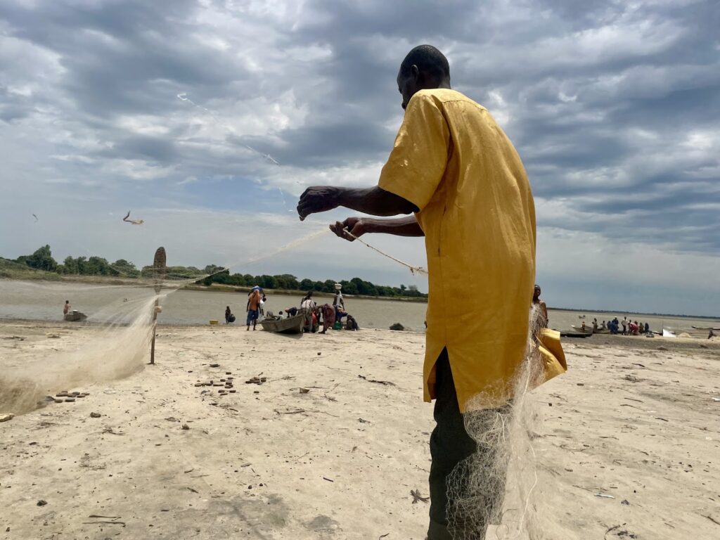 A fisherman in a yellow shirt casting a net on a sandy beach with people and boats in the background.