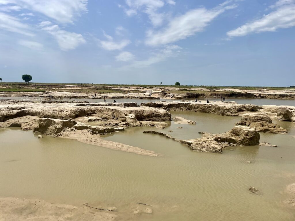 An expansive muddy landscape with standing water and eroded soil formations under a blue sky.