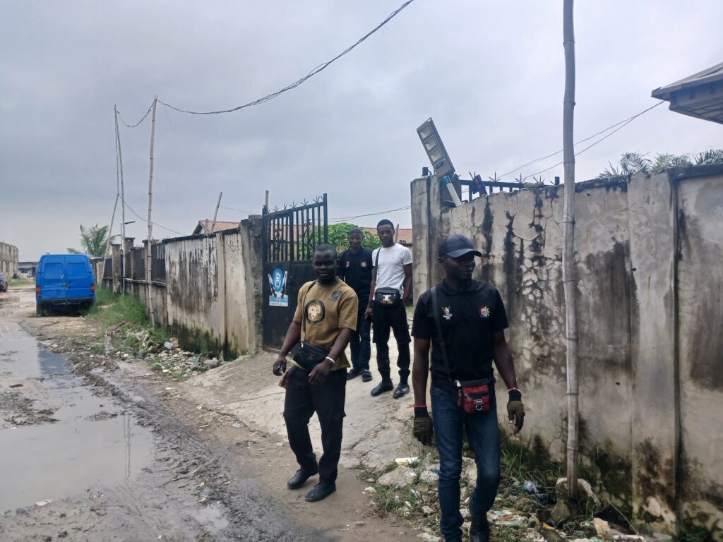 Security personnel patrolling a muddy street next to a weathered wall and a blue van.