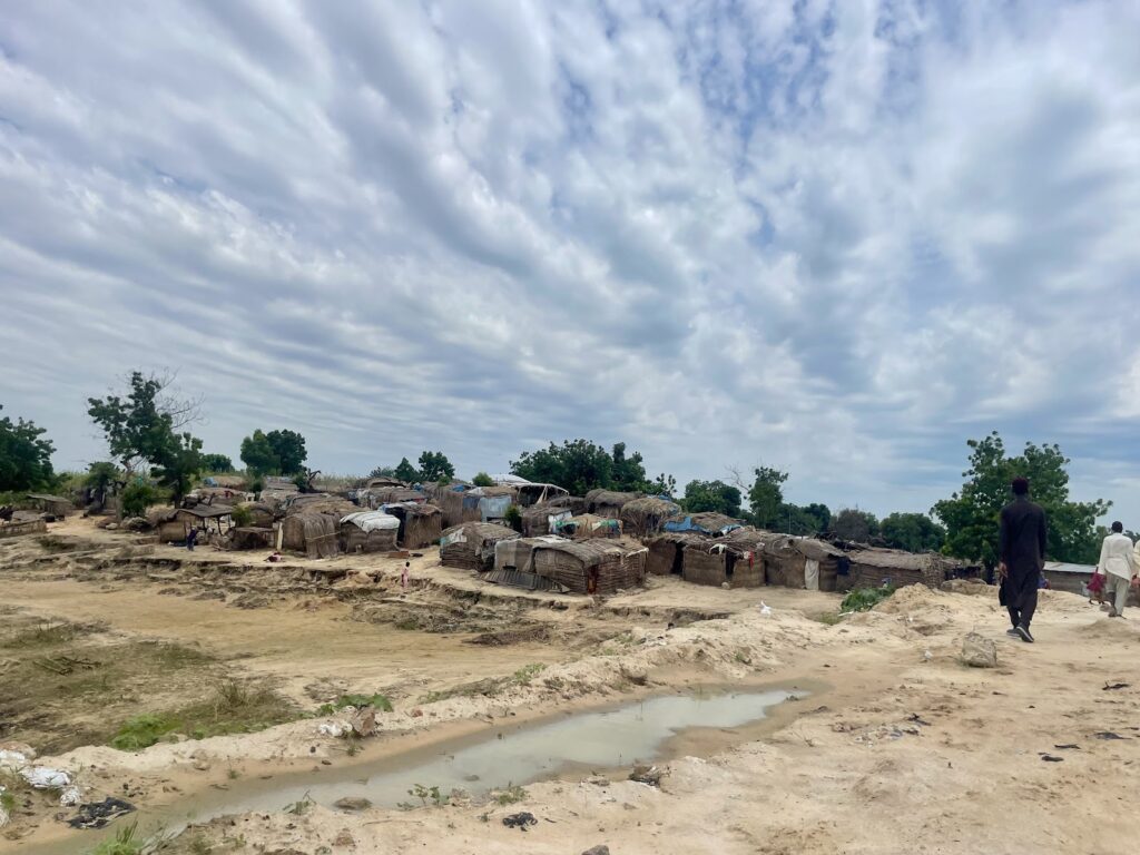 Rural landscape with makeshift huts under a cloudy sky, with people walking through the area.