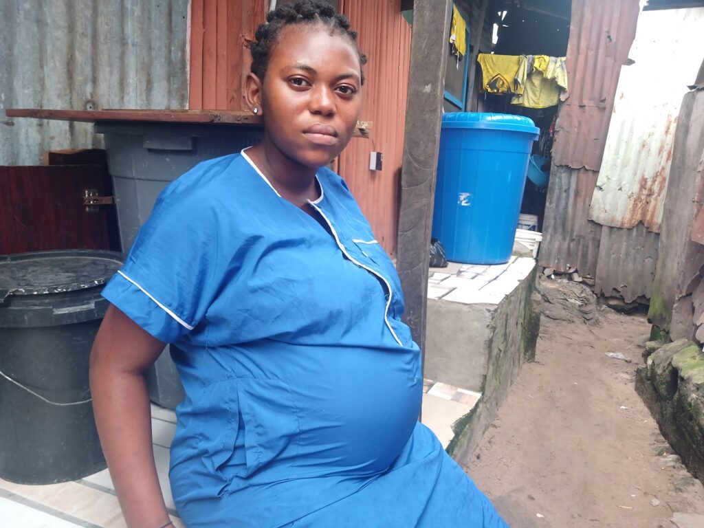 A woman in blue scrubs sitting in a narrow alleyway with a thoughtful expression.