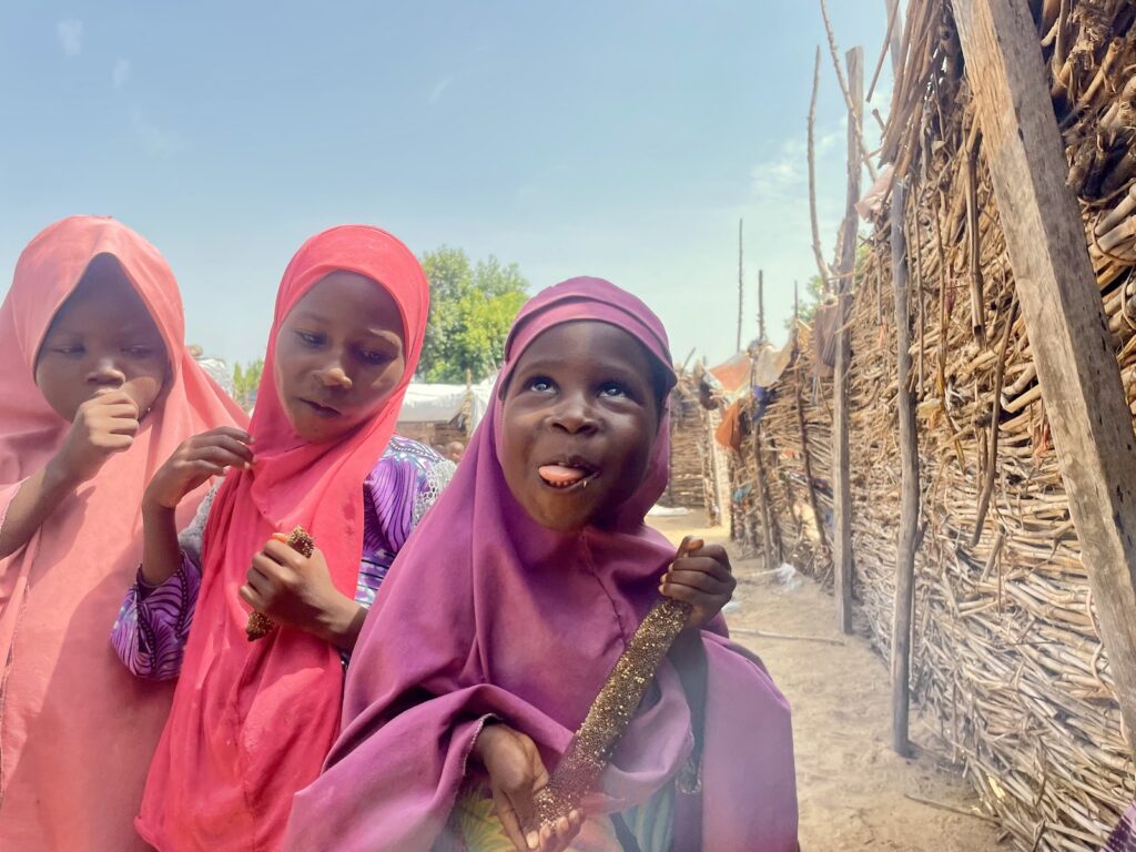 Three girls in pink headscarves, one playfully sticking out her tongue, with a rustic fence in the background.