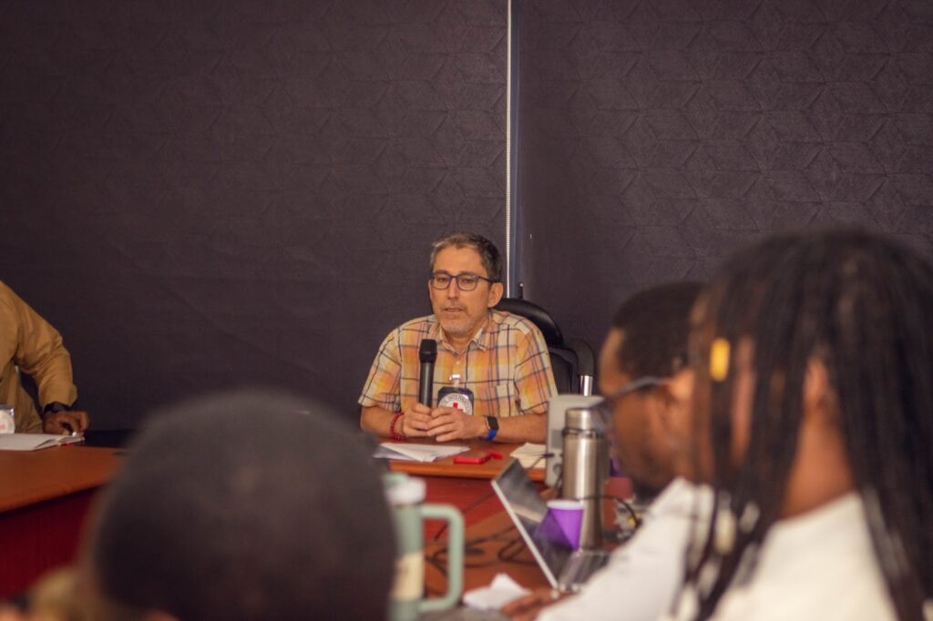 Man with glasses speaking into a microphone at a conference table with other participants out of focus.