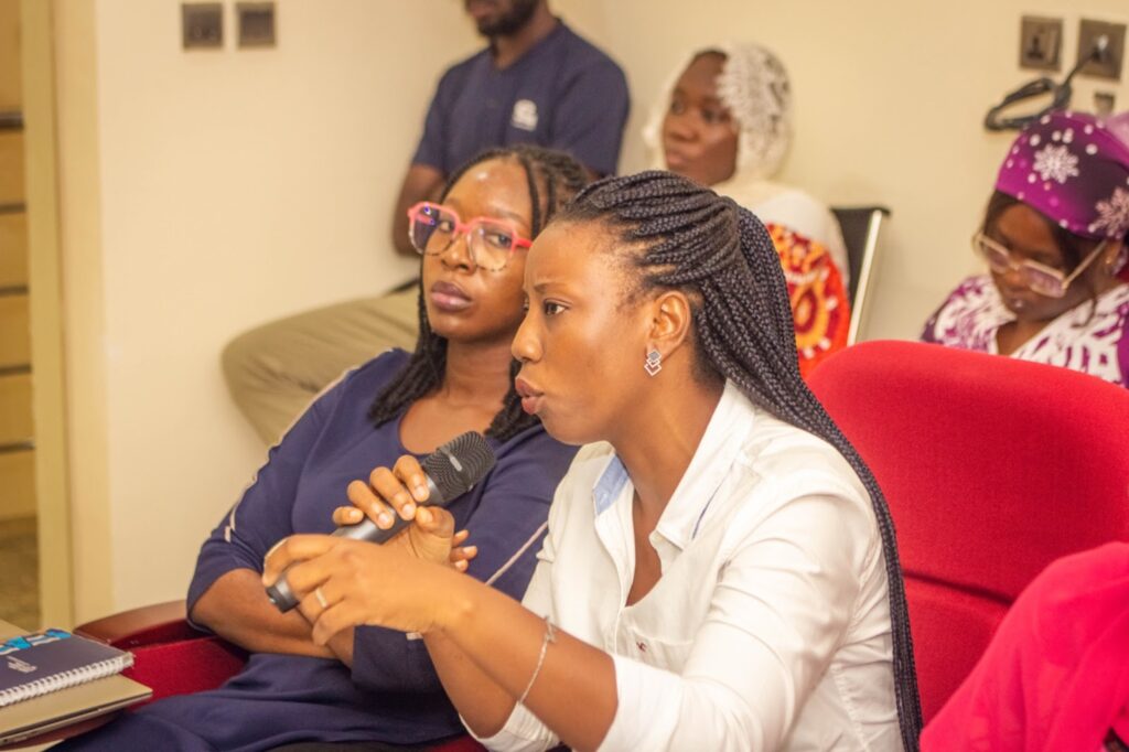 A woman speaks into a microphone at an indoor event, with attentive listeners around her.