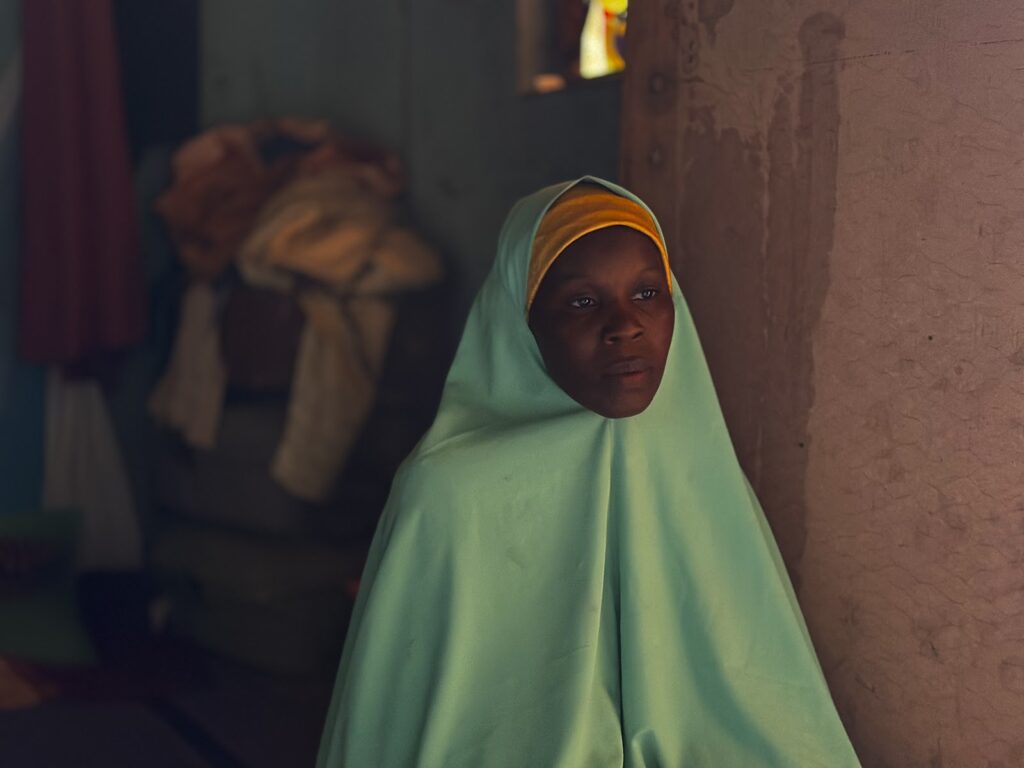 A young person in a light green and yellow shawl gazes pensively indoors, with soft lighting and a textured wall background.