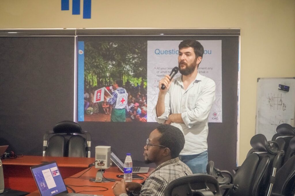 Man presenting in a conference room with attendees and a Red Cross slideshow in the background.