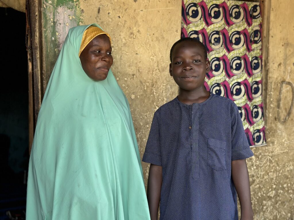 A smiling woman in a green hijab and a boy in a blue shirt standing in front of a patterned curtain.