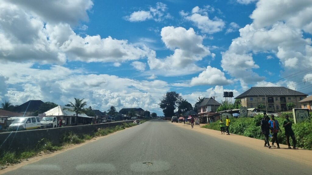 Open road in a tropical town with pedestrians, vehicles, and buildings under a bright sky with fluffy clouds.