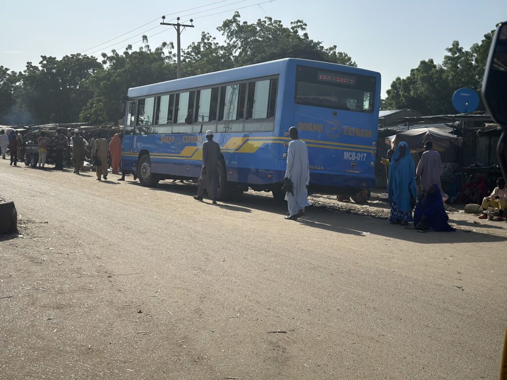 Blue and yellow Borno Express bus at a bustling market with people in colorful attire walking by.