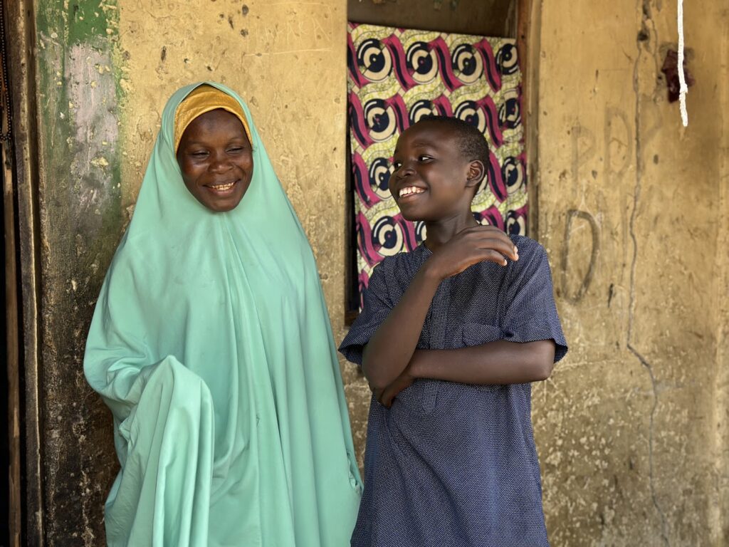 A smiling woman in a hijab beside a grinning boy against a rustic wall.