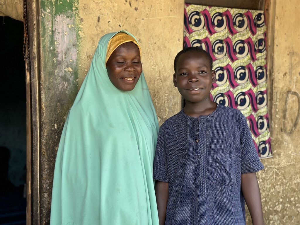 A smiling woman in a green hijab and a boy in a blue shirt standing in front of a patterned curtain.