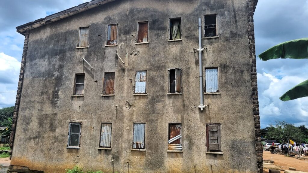 A dilapidated multi-story building with boarded-up windows, flanked by greenery under a cloudy sky.