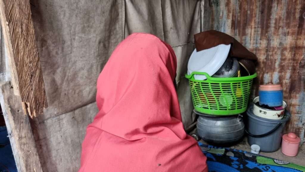 Person in a pink scarf sitting facing away, with household items and broom in the background.