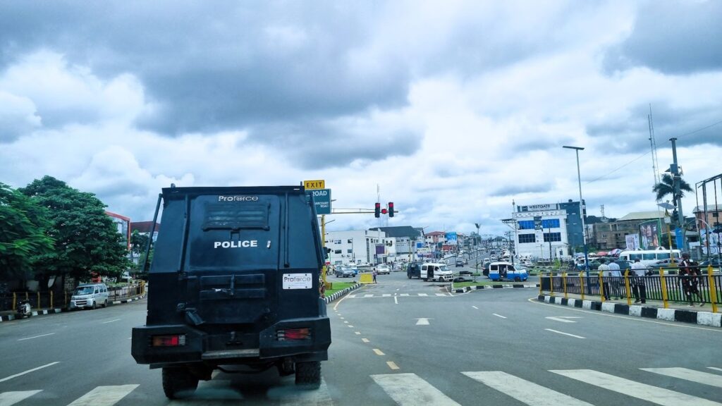 A police armored vehicle on a road with traffic lights and cityscape under cloudy skies.