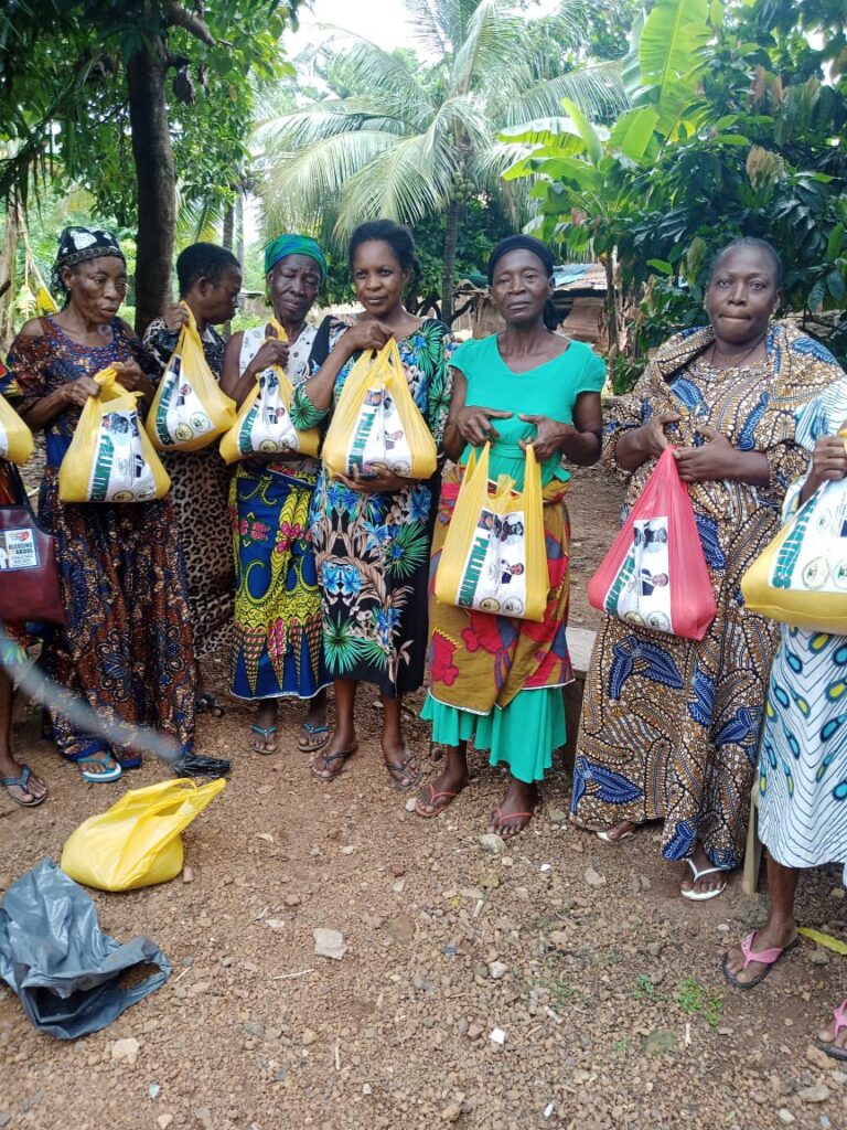 Group of women in colorful clothing holding bags, standing outdoors with greenery in the background.