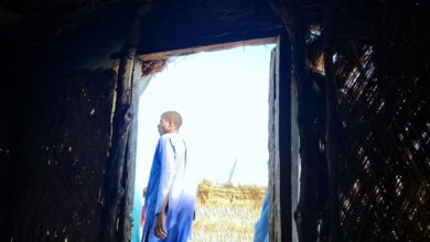 A person in blue clothing standing at the entrance of a thatched hut, looking outward.