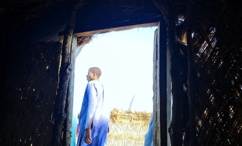 A person in blue clothing standing at the entrance of a thatched hut, looking outward.