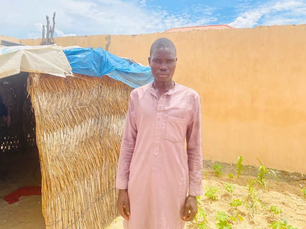 Young man in pink shirt standing in front of a thatched structure and a yellow wall under a bright sky.