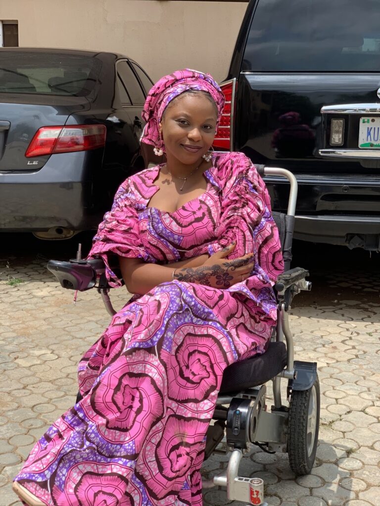 A woman in a pink patterned outfit and headscarf sitting in a wheelchair, smiling with henna on her hand.