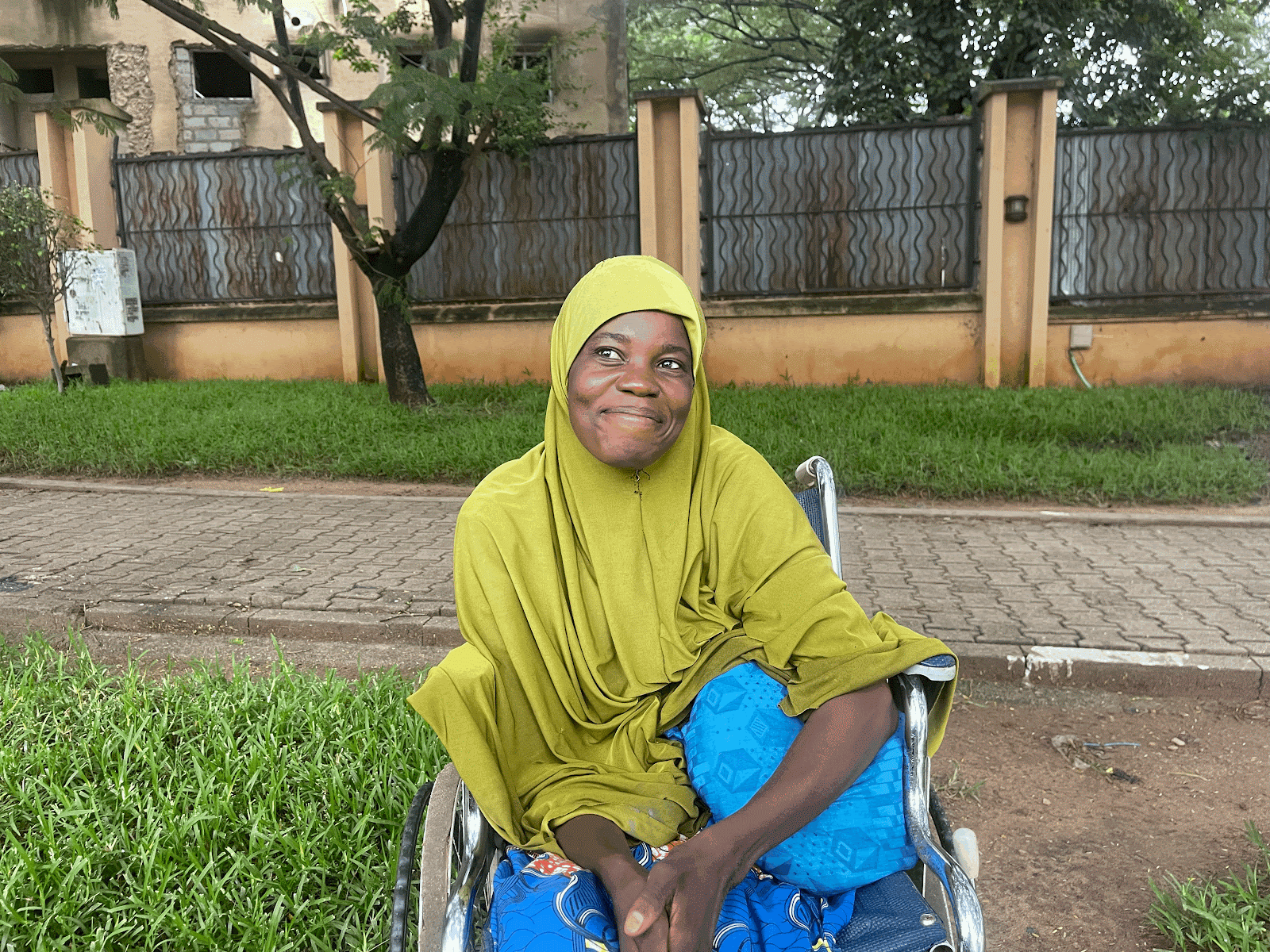 Woman in a wheelchair smiling outdoors with trees and a fence in the background.