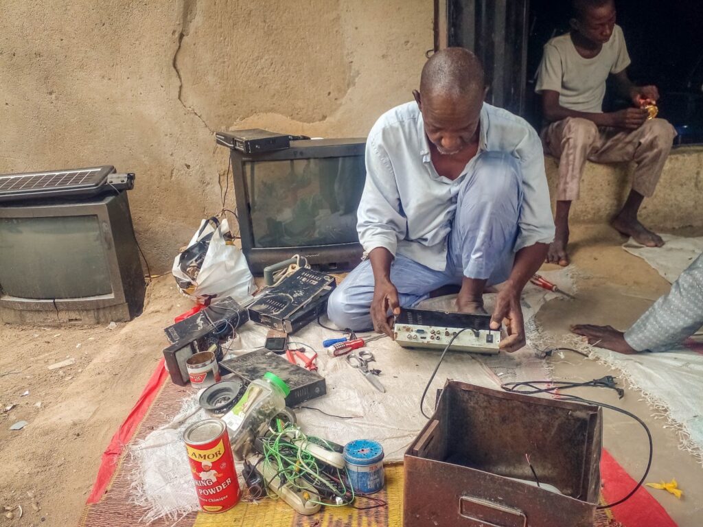 Man repairing electronics on a ground sheet with tools and old TVs around, another person watching in the background.