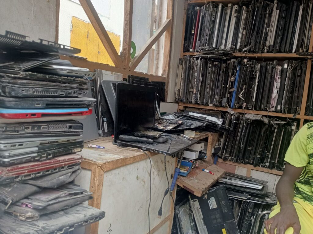 A cluttered workshop with stacks of old laptops and computer parts, and a person in a yellow top.