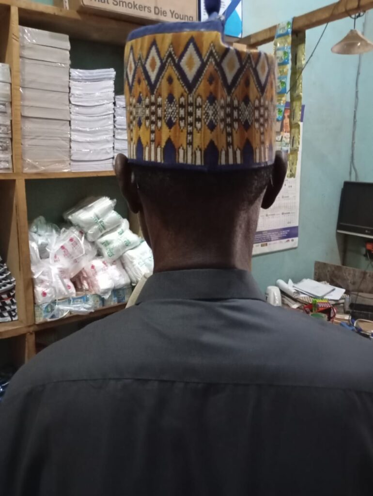 Man standing with back to camera wearing a patterned traditional cap inside a shop.