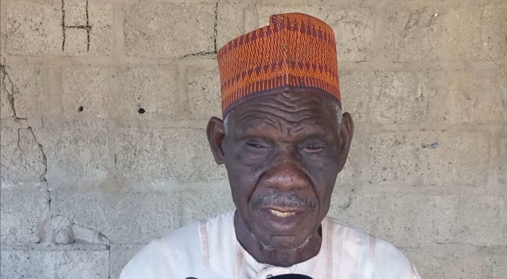 Elderly man with a weathered expression wearing a traditional hat against a concrete wall.