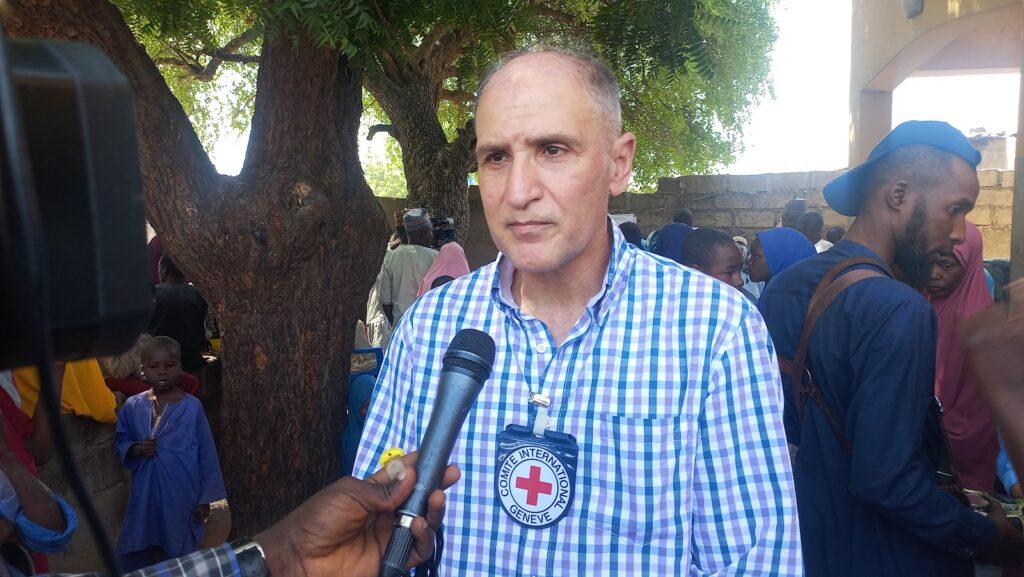 Man in checkered shirt with Red Cross badge being interviewed outdoors.