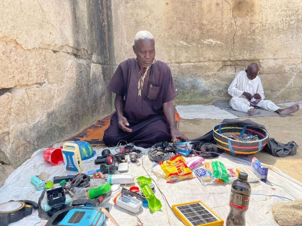 Man sitting with various items for sale, including torches and snacks, with another man in background.