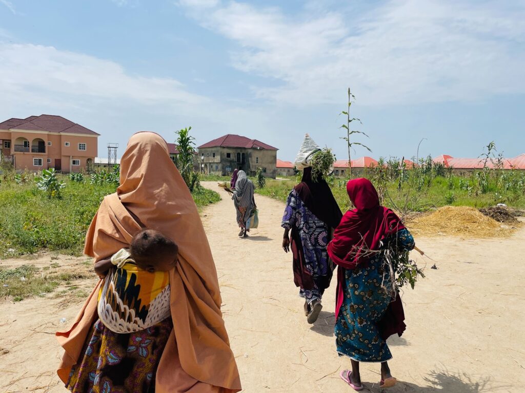 Women and a child walking on a dirt path in a developing area with buildings in the background.