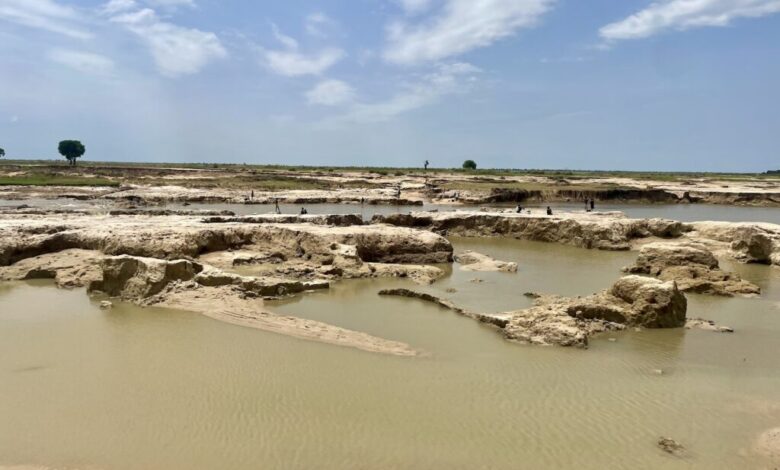 Sandy landscape with water pools under a cloudy sky, with sparse vegetation and distant figures.