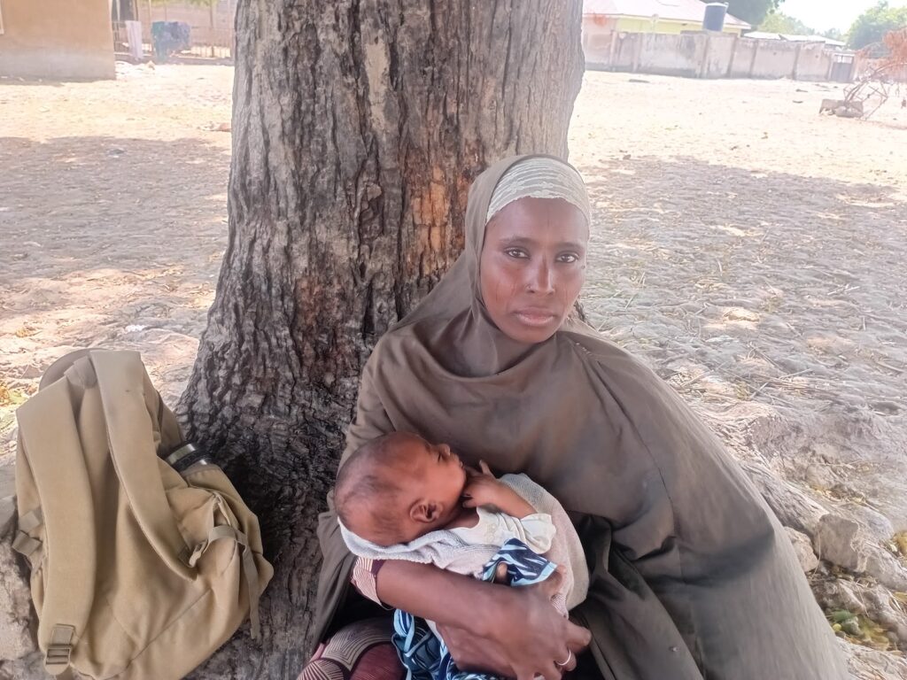 Woman sitting under a tree holding a sleeping baby, with a backpack beside her.