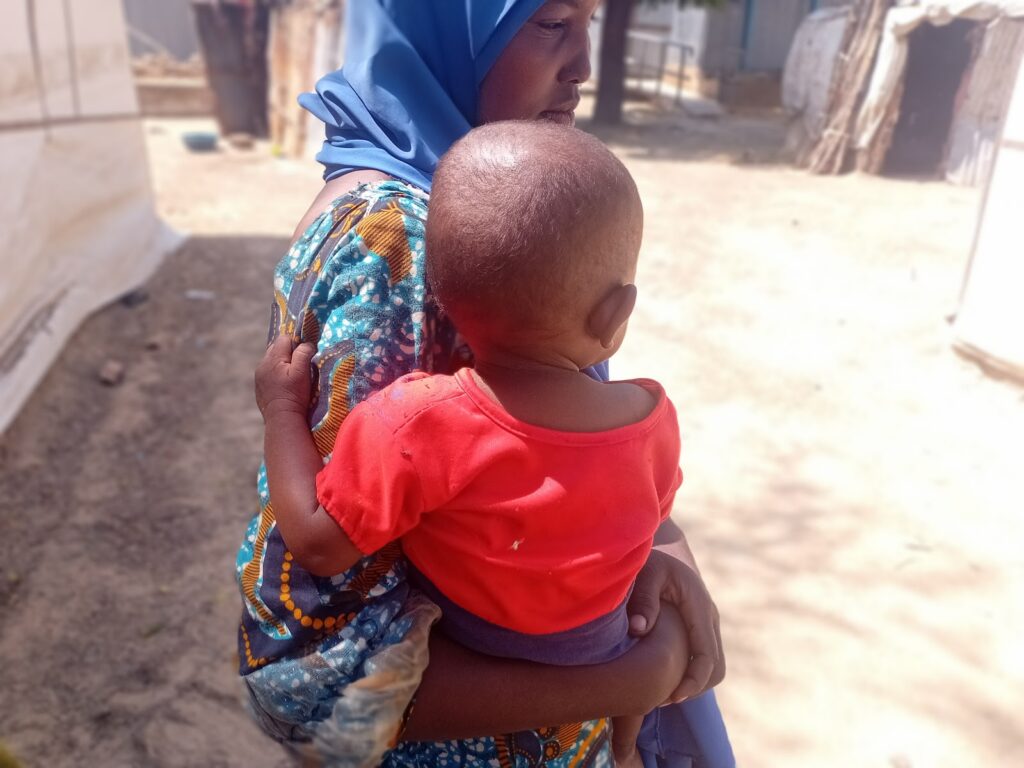 Woman in hijab holding a baby in a red shirt, standing outdoors with huts in the background.