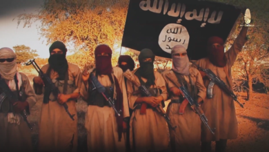 Group of armed individuals with faces covered, standing under a flag with Arabic inscription.