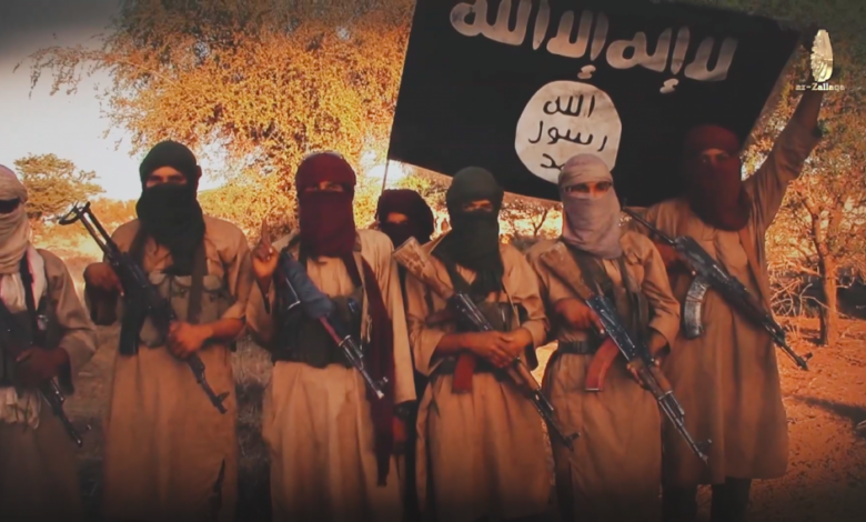 Group of armed individuals with faces covered, standing under a flag with Arabic inscription.