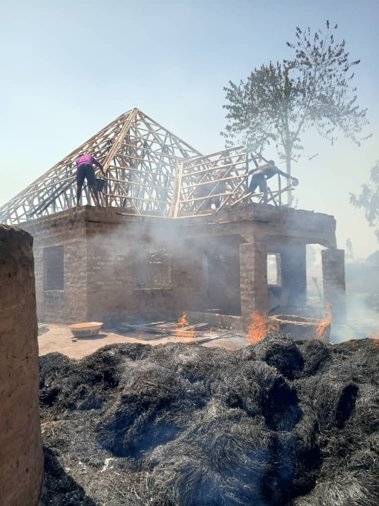 Workers build a wooden roof structure atop a stone building with smoldering ashes in the foreground.