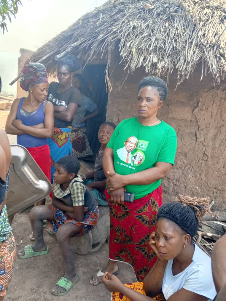 Group of people gathered outside a thatched hut with various expressions.