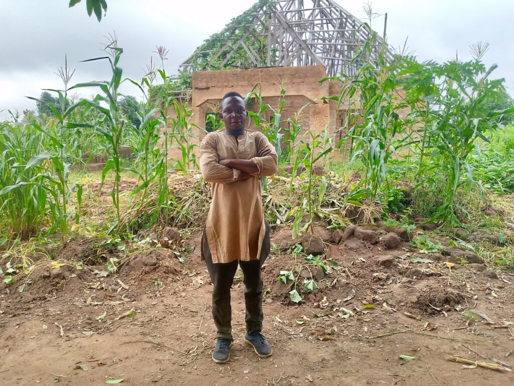 Man standing with arms crossed in front of a building under construction amidst green foliage.