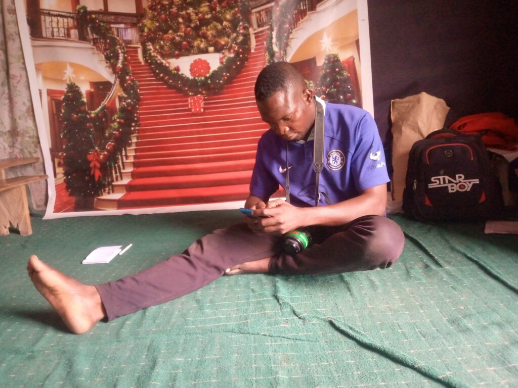 Man in blue jersey sits on floor, tuning a camera, with festive backdrop and backpack nearby.